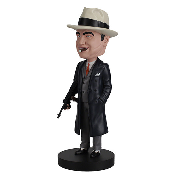 3/4 left side view of Al Capone bobblehead, wearing a white fedora, dark trench coat, and suit, holds a gun with a smirk. The base is labeled "Al Capone." Retro and playful tone.