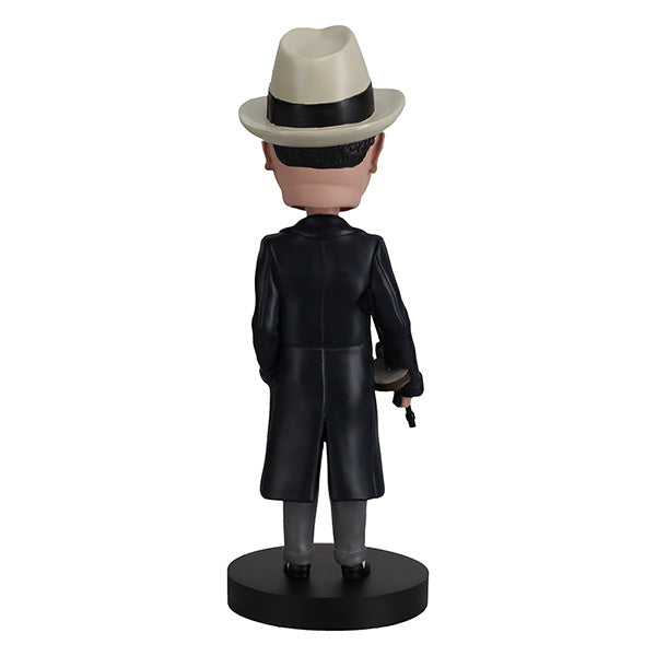 Back view of Al Capone bobblehead, wearing a white fedora, dark trench coat, and suit, holds a gun with a smirk. The base is labeled "Al Capone." Retro and playful tone.