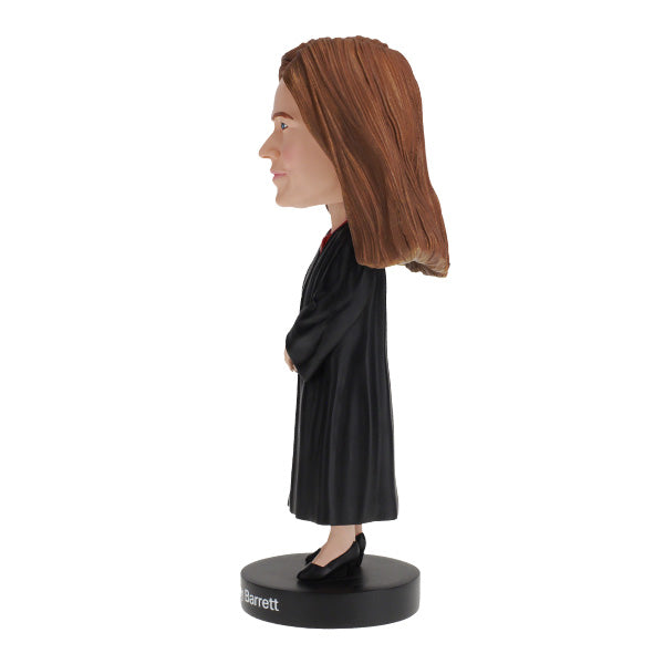 Left side view of Amy Coney Barrett Bobblehead figure is wearing judicial robes and long brown hair, standing on a base labeled "Amy Coney Barrett," conveying authority.