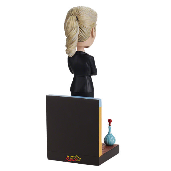 Back view of Better Call Saul Kim Wexler Bobblehead features a woman in a black suit with arms crossed, standing confidently in front of a chart backdrop. A blue bottle with a red cap is at her feet.