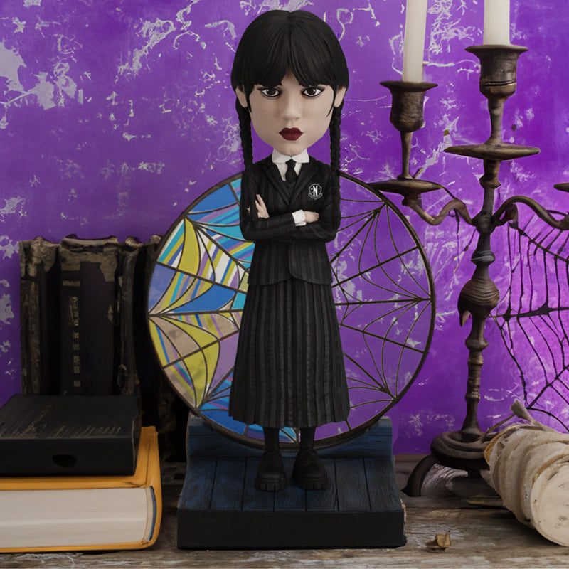 The Wednesday bobblehead shown in front of a purple background showcasing the transparent stained glass window behind her.