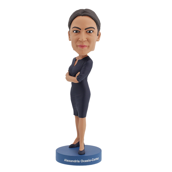 Alexandria Ocasio-Cortes bobblehead figure stands on a blue base, wearing a dark dress with arms crossed. The expression is confident and poised, conveying determination.