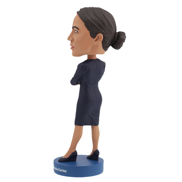 Left side view of Alexandria Ocasio-Cortes bobblehead figure stands on a blue base, wearing a dark dress with arms crossed. The expression is confident and poised, conveying determination.