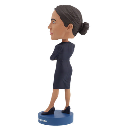 Left side view of Alexandria Ocasio-Cortes bobblehead figure stands on a blue base, wearing a dark dress with arms crossed. The expression is confident and poised, conveying determination.