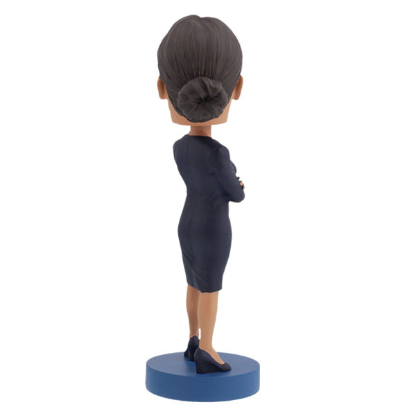 Back view of Alexandria Ocasio-Cortes bobblehead figure stands on a blue base, wearing a dark dress with arms crossed. The expression is confident and poised, conveying determination.