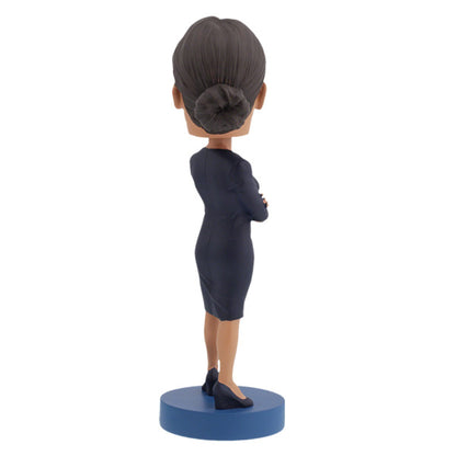 Back view of Alexandria Ocasio-Cortes bobblehead figure stands on a blue base, wearing a dark dress with arms crossed. The expression is confident and poised, conveying determination.