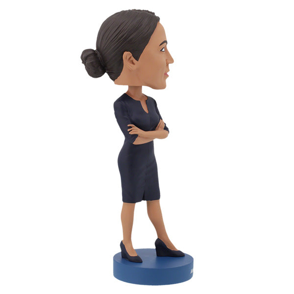 Right side view of Alexandria Ocasio-Cortes bobblehead figure stands on a blue base, wearing a dark dress with arms crossed. The expression is confident and poised, conveying determination.
