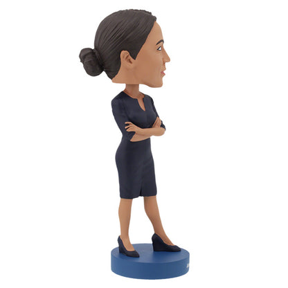 Right side view of Alexandria Ocasio-Cortes bobblehead figure stands on a blue base, wearing a dark dress with arms crossed. The expression is confident and poised, conveying determination.