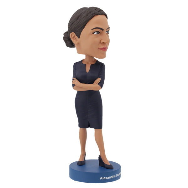 3/4 right side view of Alexandria Ocasio-Cortes bobblehead figure stands on a blue base, wearing a dark dress with arms crossed. The expression is confident and poised, conveying determination.