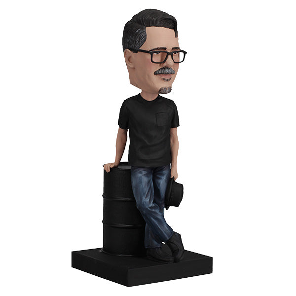 3/4 right view Vince Gilligan bobblehead with glasses and mustache holding hat, leaning on barrel on green base