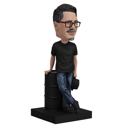 3/4 right view Vince Gilligan bobblehead with glasses and mustache holding hat, leaning on barrel on green base