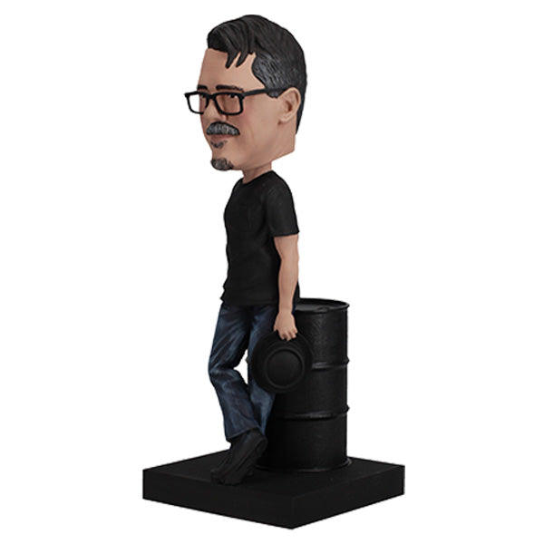 3/4 View of Vince Gilligan with glasses and mustache holding hat, leaning on barrel on green base