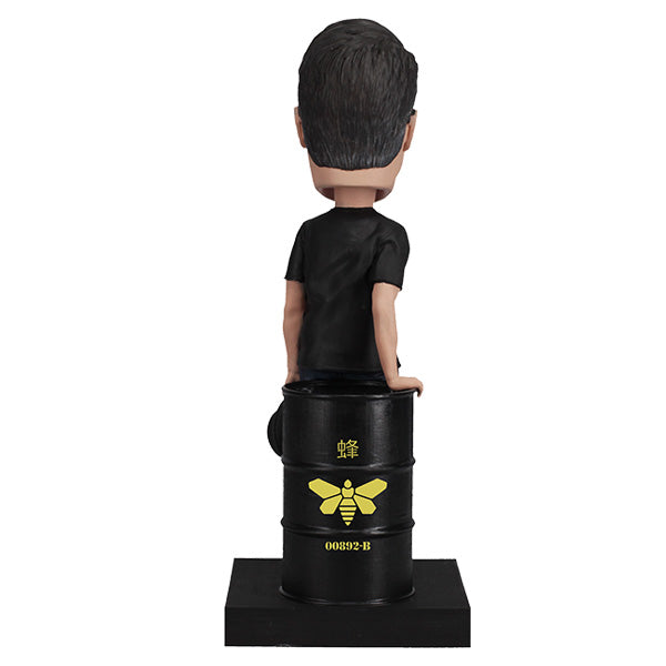 Back view Vince Gilligan bobblehead with glasses and mustache holding hat, leaning on barrel on green base