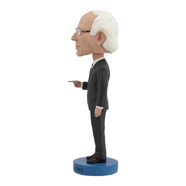 Left side view of Bernie Sanders Bobblehead figurine of an elderly man with white hair and glasses, wearing a dark suit, standing on a base labeled "Bernie Sanders." The figure has a calm expression.