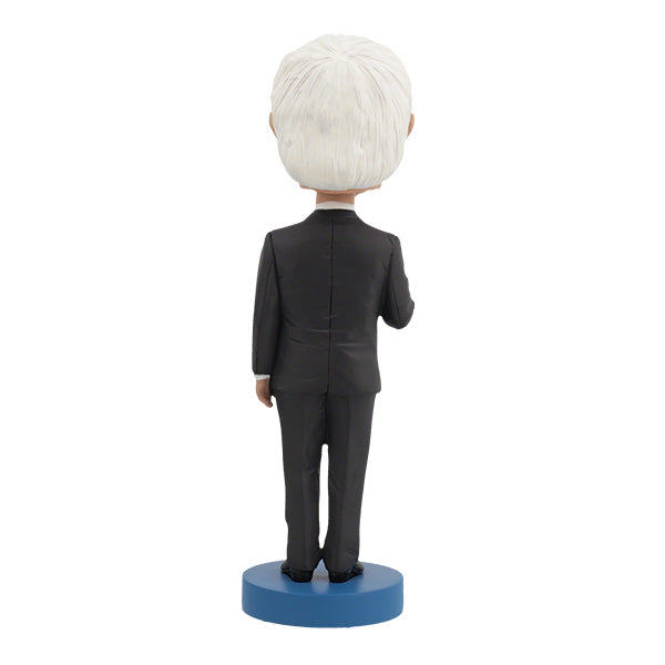 Back View of Bernie Sanders Bobblehead figurine of an elderly man with white hair and glasses, wearing a dark suit, standing on a base labeled "Bernie Sanders." The figure has a calm expression.