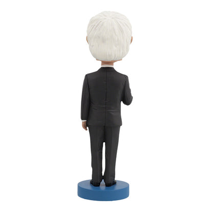 Back View of Bernie Sanders Bobblehead figurine of an elderly man with white hair and glasses, wearing a dark suit, standing on a base labeled "Bernie Sanders." The figure has a calm expression.