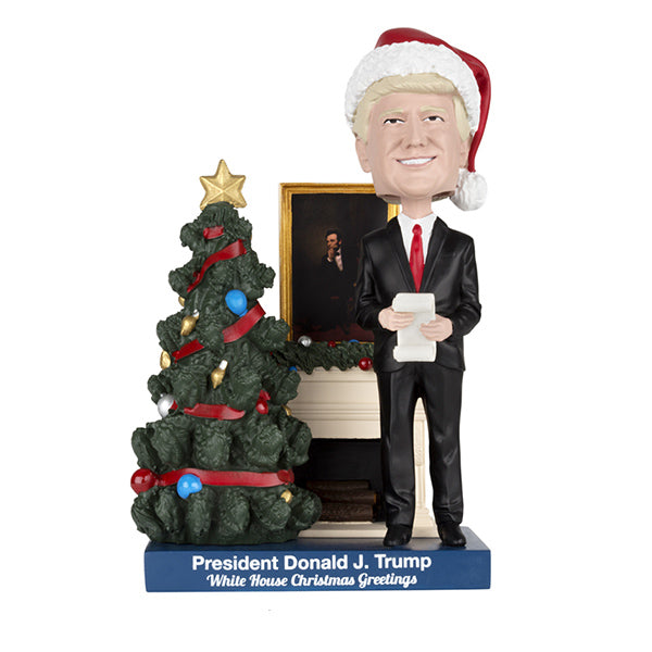 Donald J. Trump bobblehead in suit and Santa hat beside Christmas tree with holiday greeting text.
