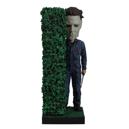 Masked figure in blue jumpsuit holding knife behind hedge.