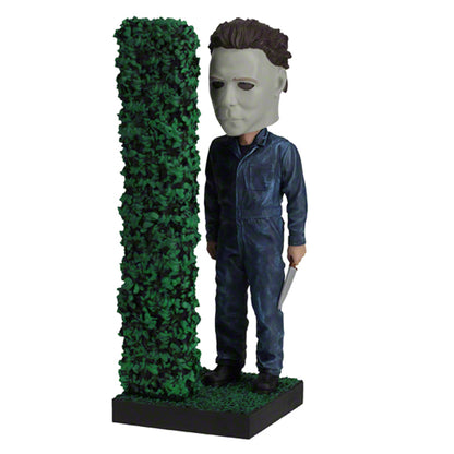 3/4 left view Masked figure in blue jumpsuit holding knife behind hedge.