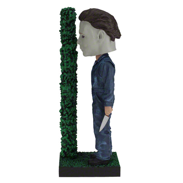 Left view Masked figure in blue jumpsuit holding knife behind hedge.
