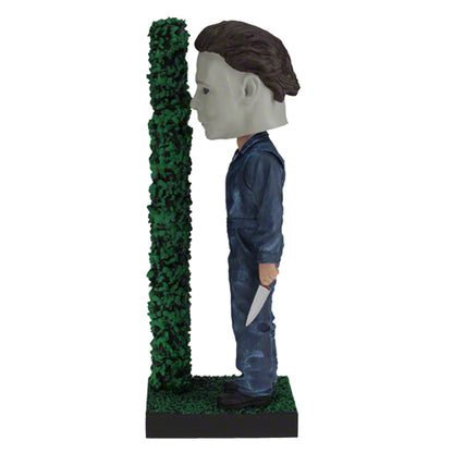 Left view Masked figure in blue jumpsuit holding knife behind hedge.