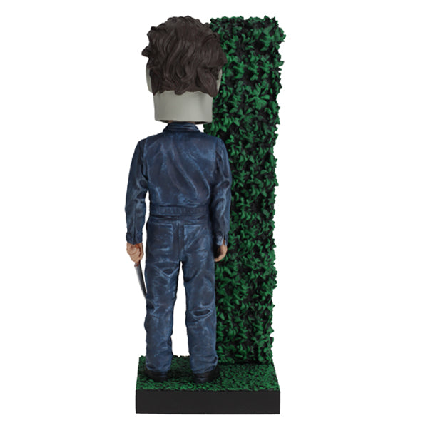Back view Masked figure in blue jumpsuit holding knife behind hedge.