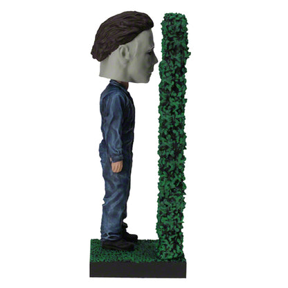 Right view masked figure in blue jumpsuit holding knife behind hedge.