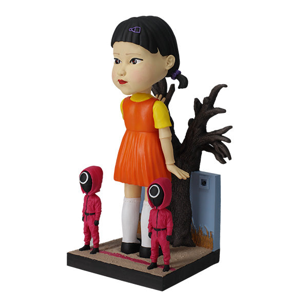 Left view large doll statue in orange and yellow dress with two small pink-hooded figures and leafless tree.