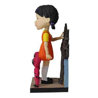 Left side large doll statue in orange and yellow dress with two small pink-hooded figures and leafless tree.