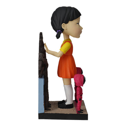 Right view large doll statue in orange and yellow dress with two small pink-hooded figures and leafless tree.