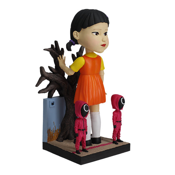 Right view large doll statue in orange and yellow dress with two small pink-hooded figures and leafless tree.
