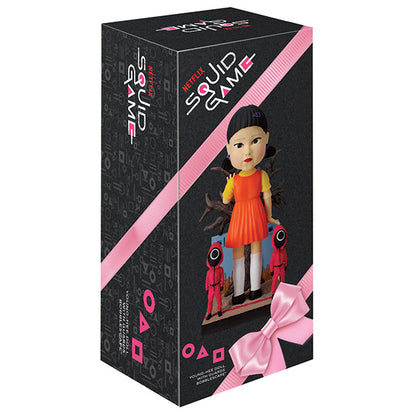 Squid Game doll box with orange-dressed doll and two masked figures on black background.